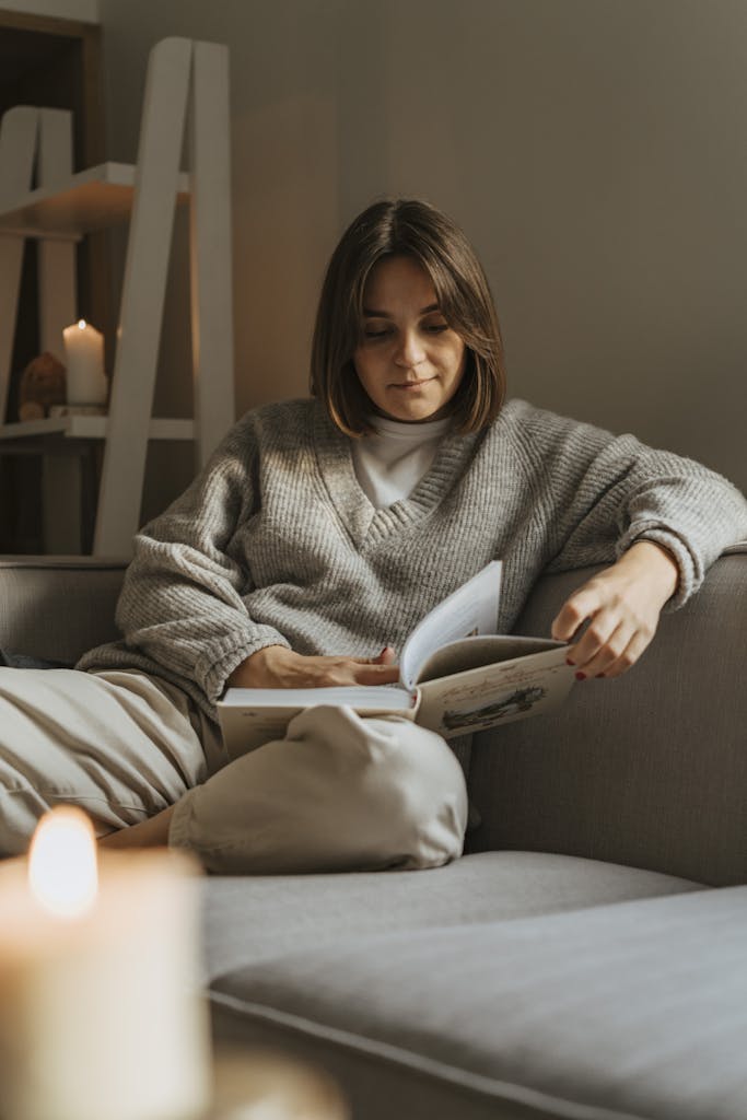 Woman in a cozy sweater reading a book on a comfortable sofa, creating a warm, serene atmosphere.